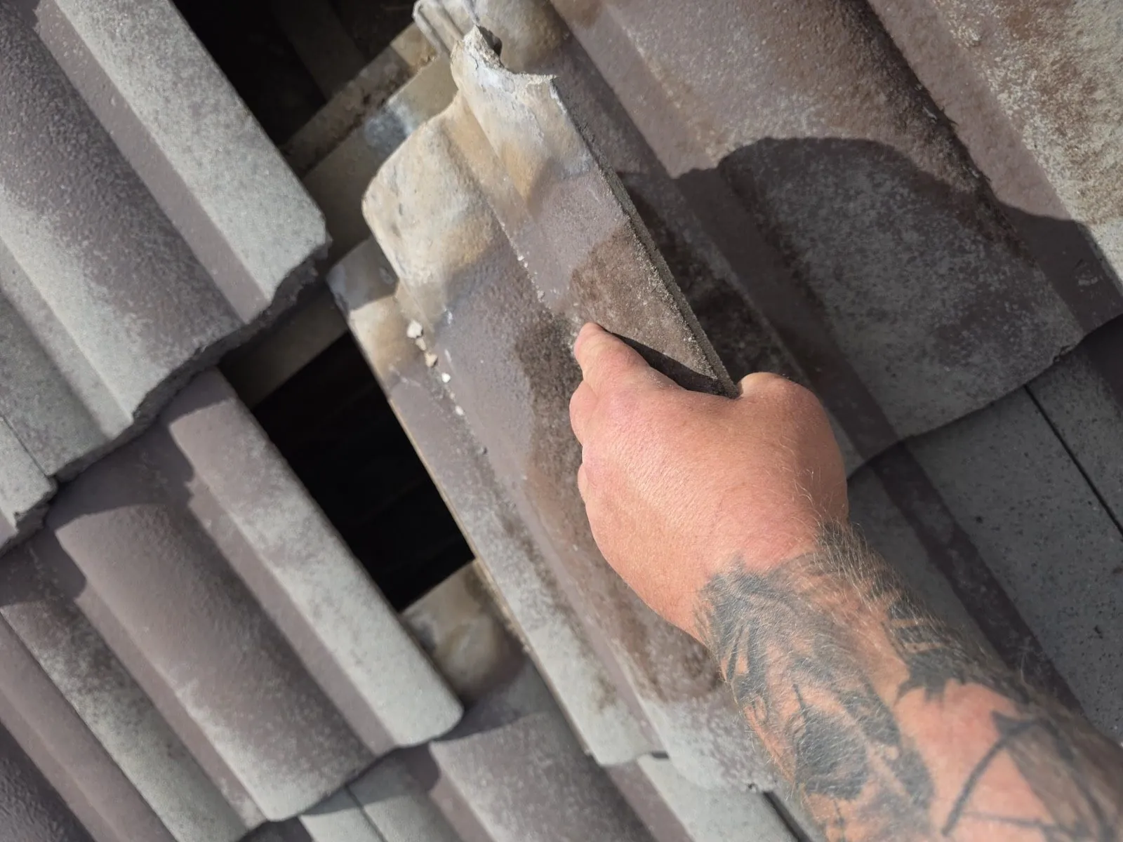 Roofer's hand lifting a cracked concrete tile clear of the roof, the timber batten and dark cavity visible in the gap left behind