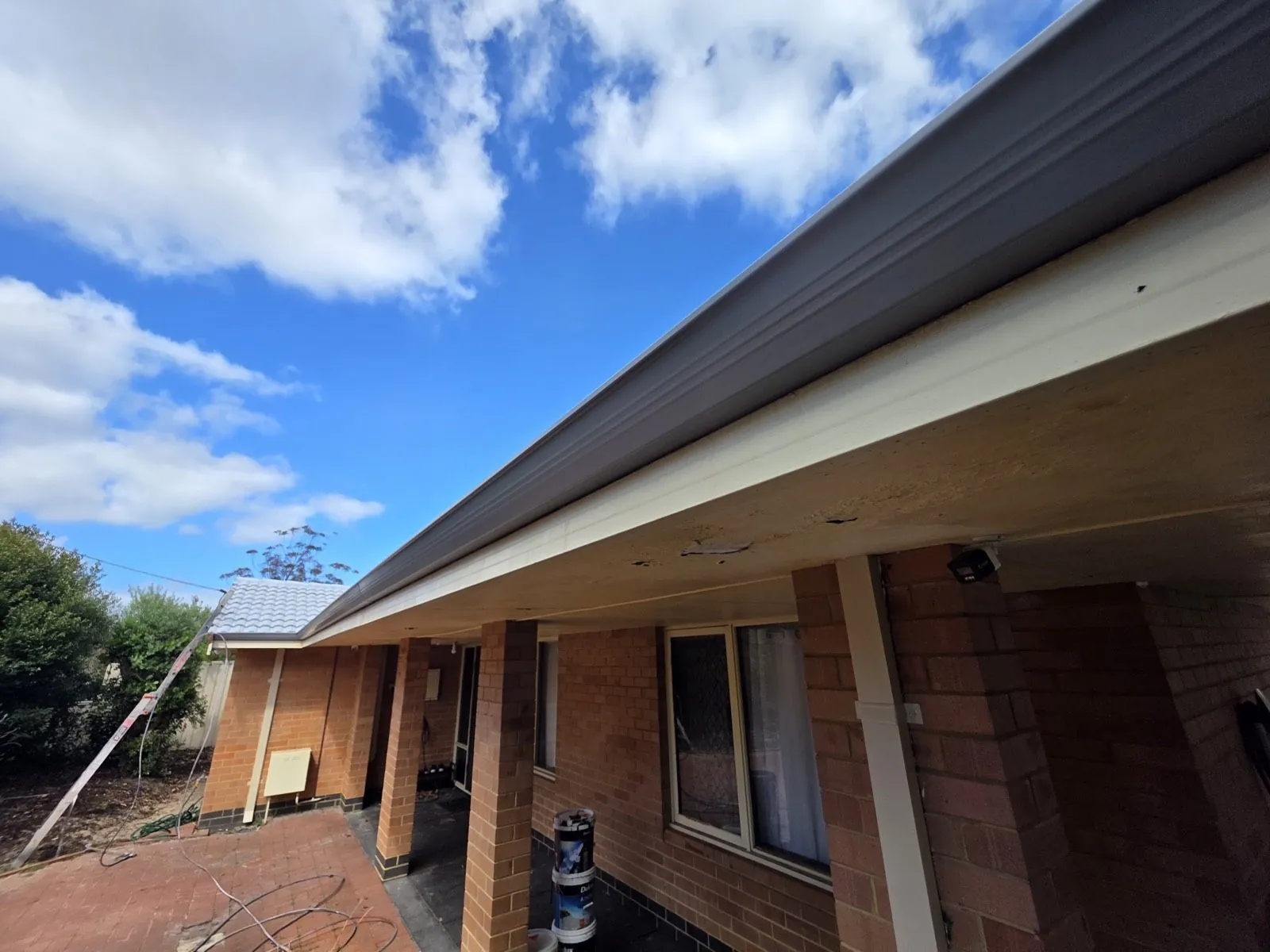 New guttering running the length of the eaves on a red brick Perth home, ladder against the wall and coating tins on the paving below