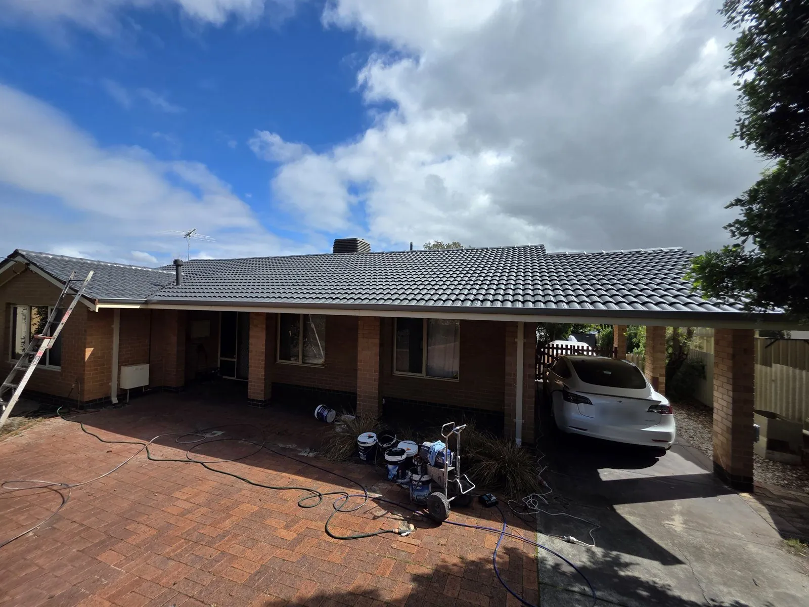 Finished charcoal tile roof on a red brick Perth home seen from the driveway, new gutters along the eaves, coating tins and spray rig still on the paving and a car parked in the carport